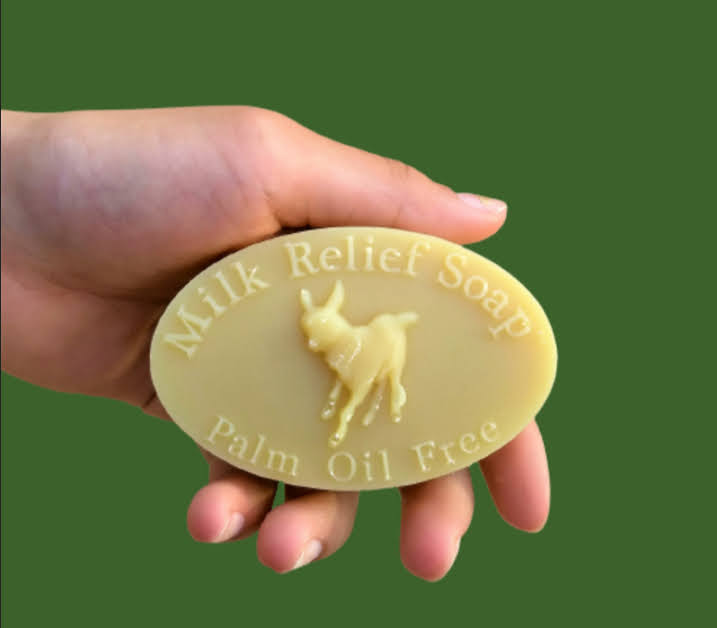 A hand holding a bar of Milk Relief Soap, featuring a design with the words 'Milk Relief Soap' and 'Palm Oil Free' along with a goat figure.